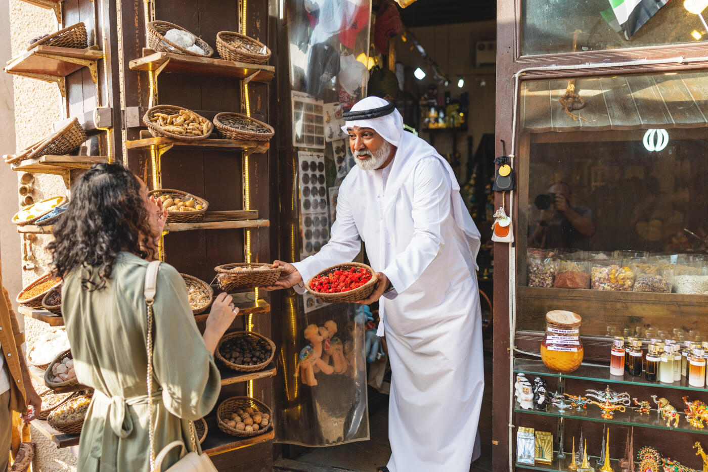A Middle Eastern woman in a green dress interacts with a local shopkeeper in a bustling outdoor spice market in Old Dubai, surrounded by vibrant spices and traditional decor under warm daylight.