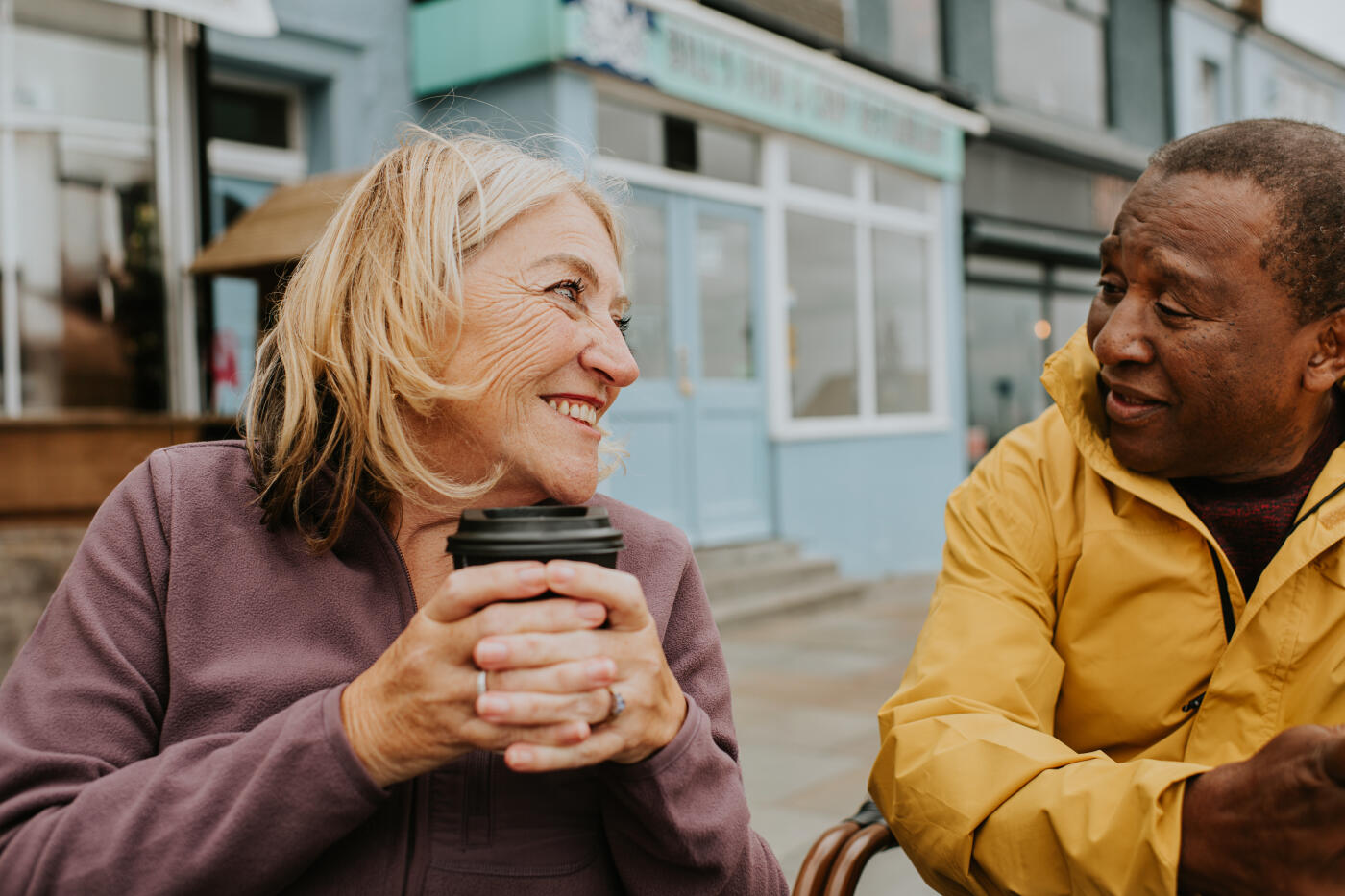 A senior couple drink coffee outside a coffee shop. Focus is on a carefree, content older woman, who smiles towards her husband and sips on a hot drink.