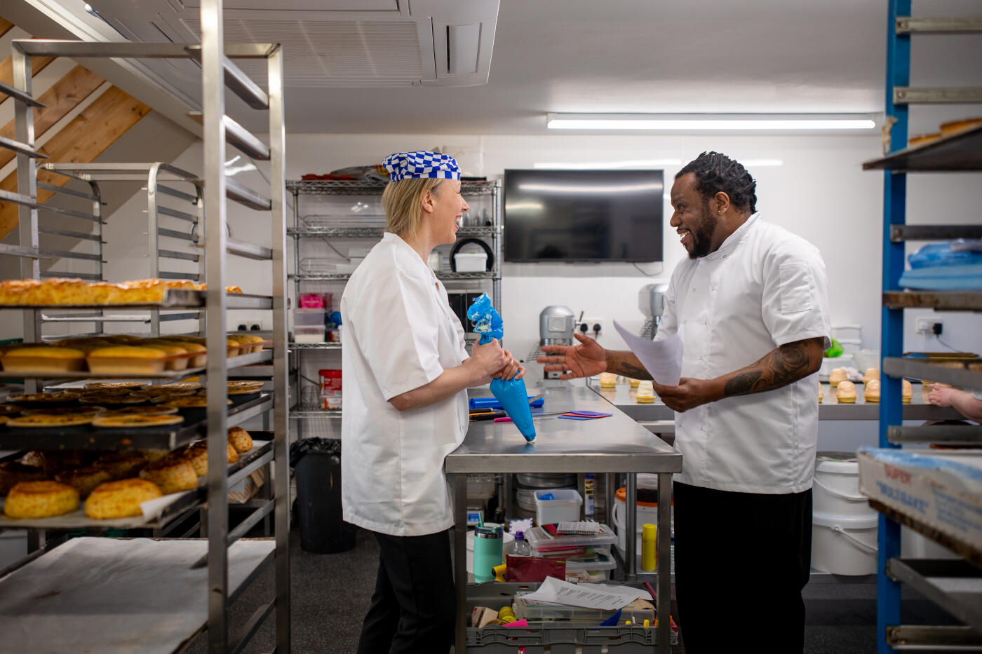 A medium shot of two male and female chefs discussing in a Northumberland bakery kitchen. One chef is holding a piping bag, and the other chef is holding a paper document. They are wearing chef's whites.Videos similar to this scenario are available.