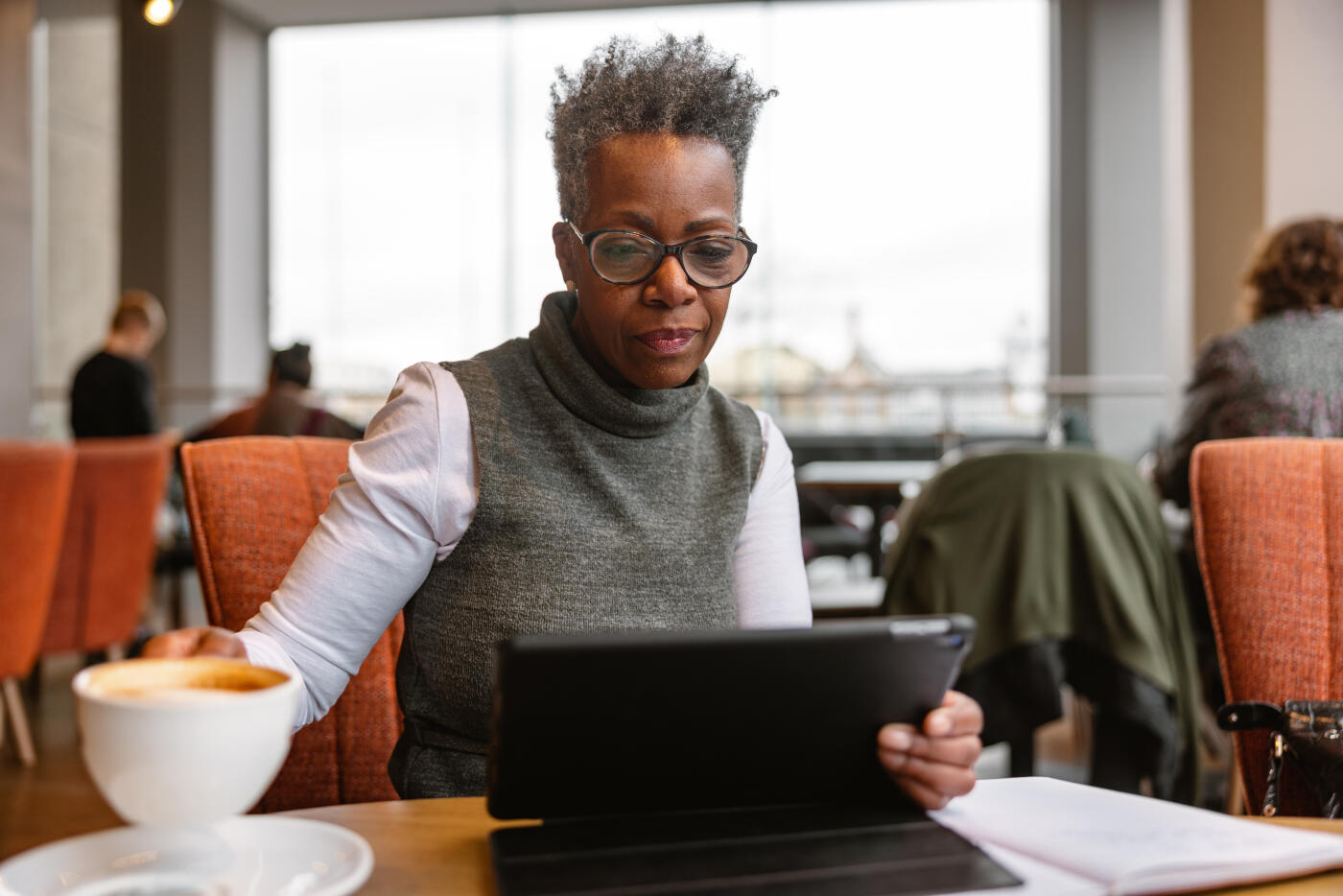 A senior adult black female business owner drinking coffee at a cafe while working remotely. She is reading the reports on the digital tablet in front of her while holding a cup of coffee in her hands. She looks focused and concerned.
