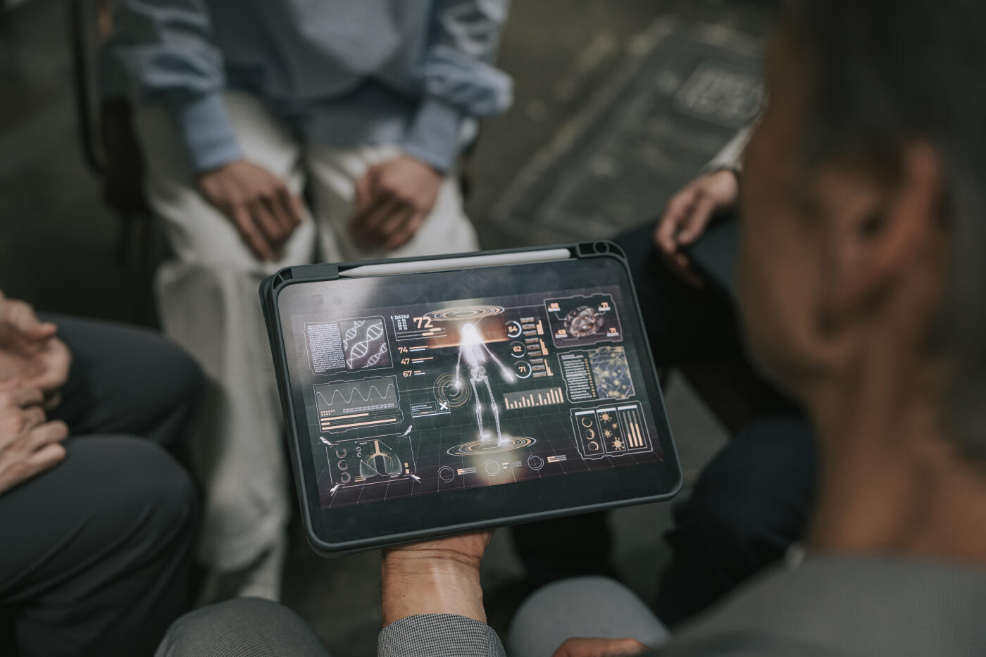 Close-up of a senior Japanese man holding a digital tablet displaying a futuristic medical interface, surrounded by a group of Japanese individuals seated in a circle. The tablet screen shows anatomical diagrams, DNA data, and health analytics. The setting suggests a collaborative discussion on healthcare innovation, medical research, or AI-driven diagnostics in a modern Japanese environment.
