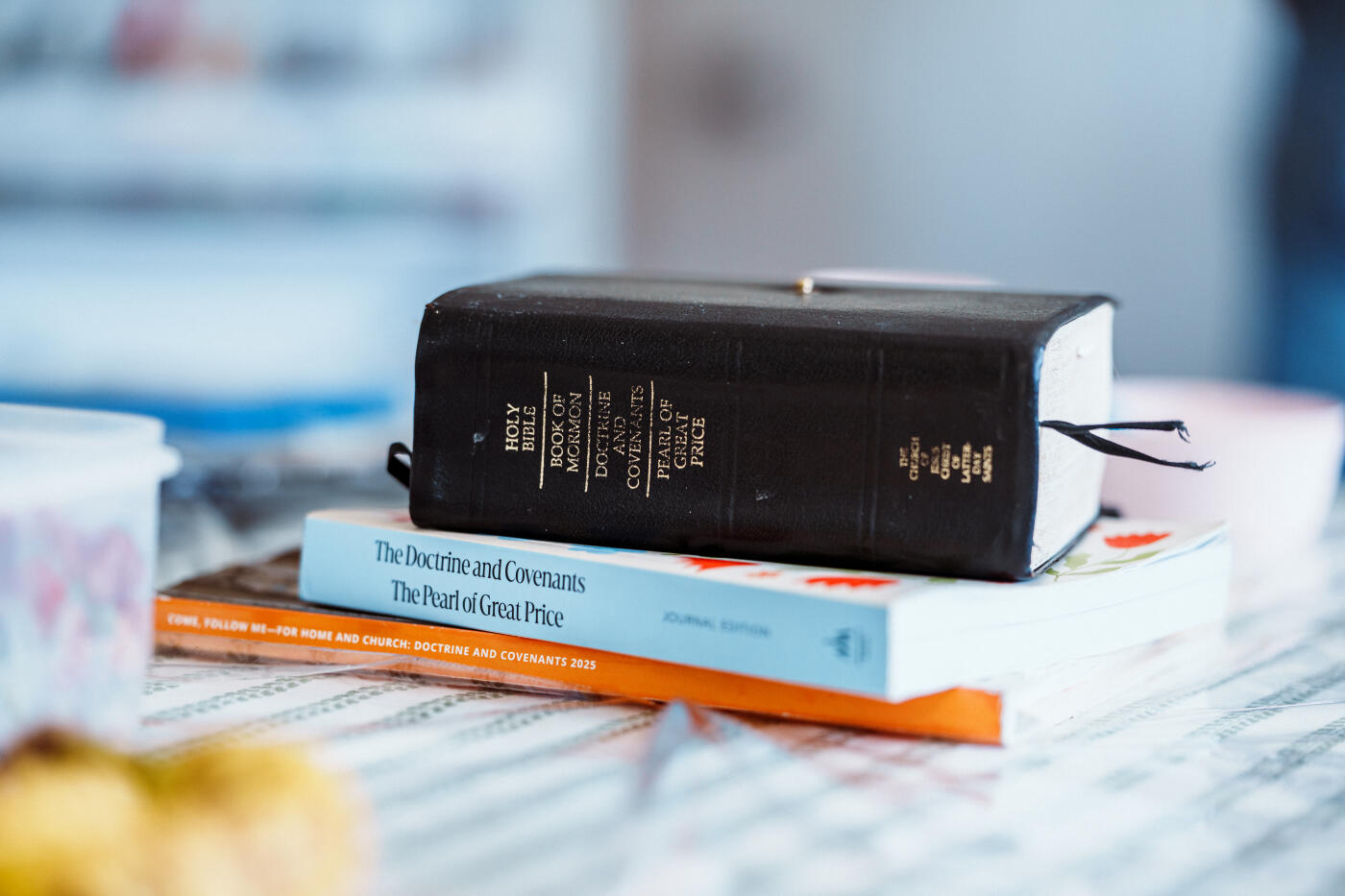 A stack of religious study books, including the Holy Bible and Book of Mormon, sits on a patterned tablecloth in a bright kitchen.