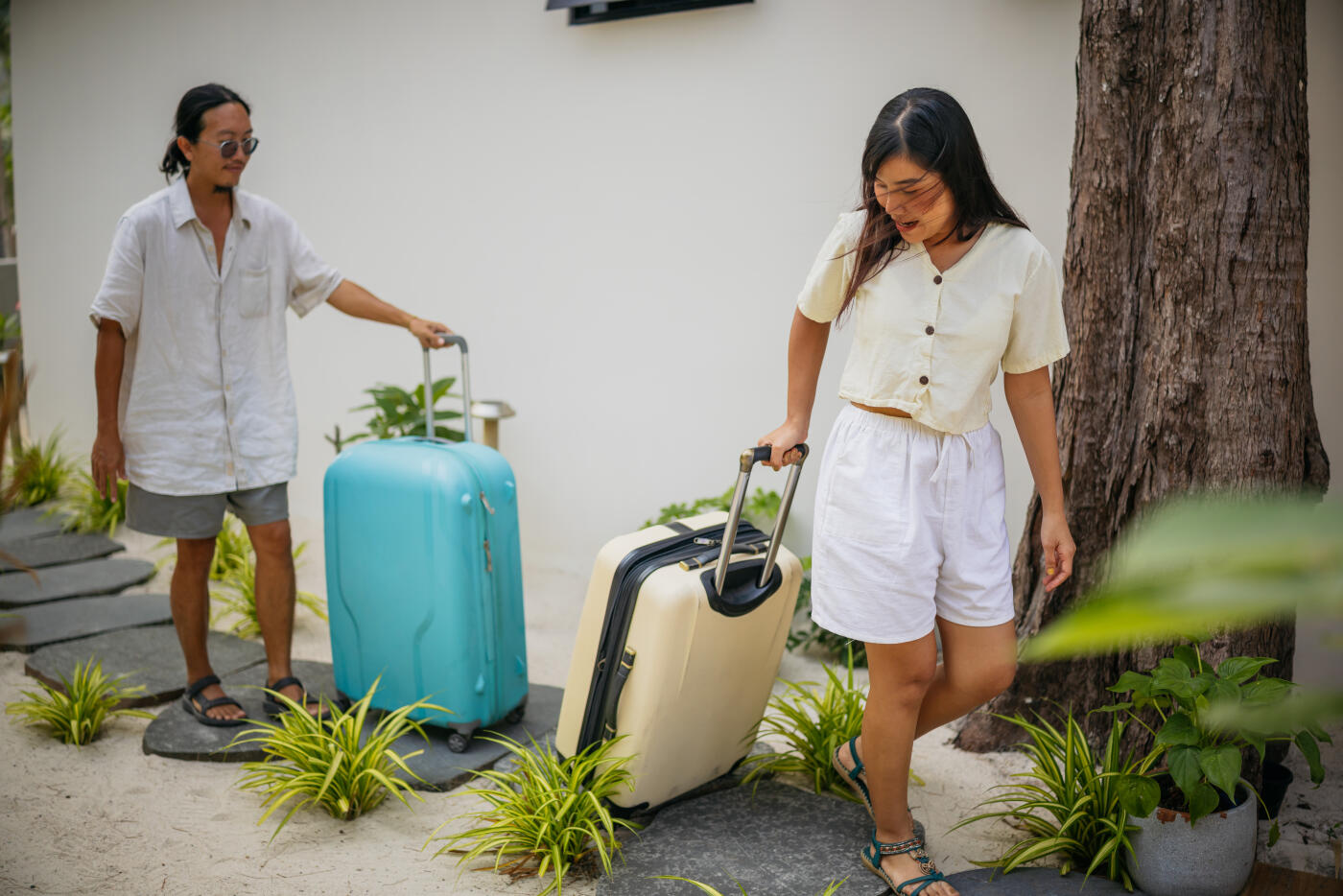 Asian couple arriving at their tropical bungalow for vacation pulling suitcases on a stone path