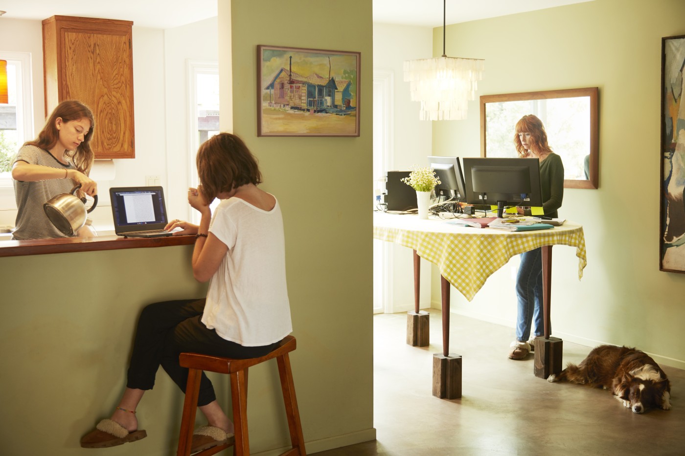 Two college age sisters are at home drinking tea in the morning with their mother in the background working from home at the kitchen table