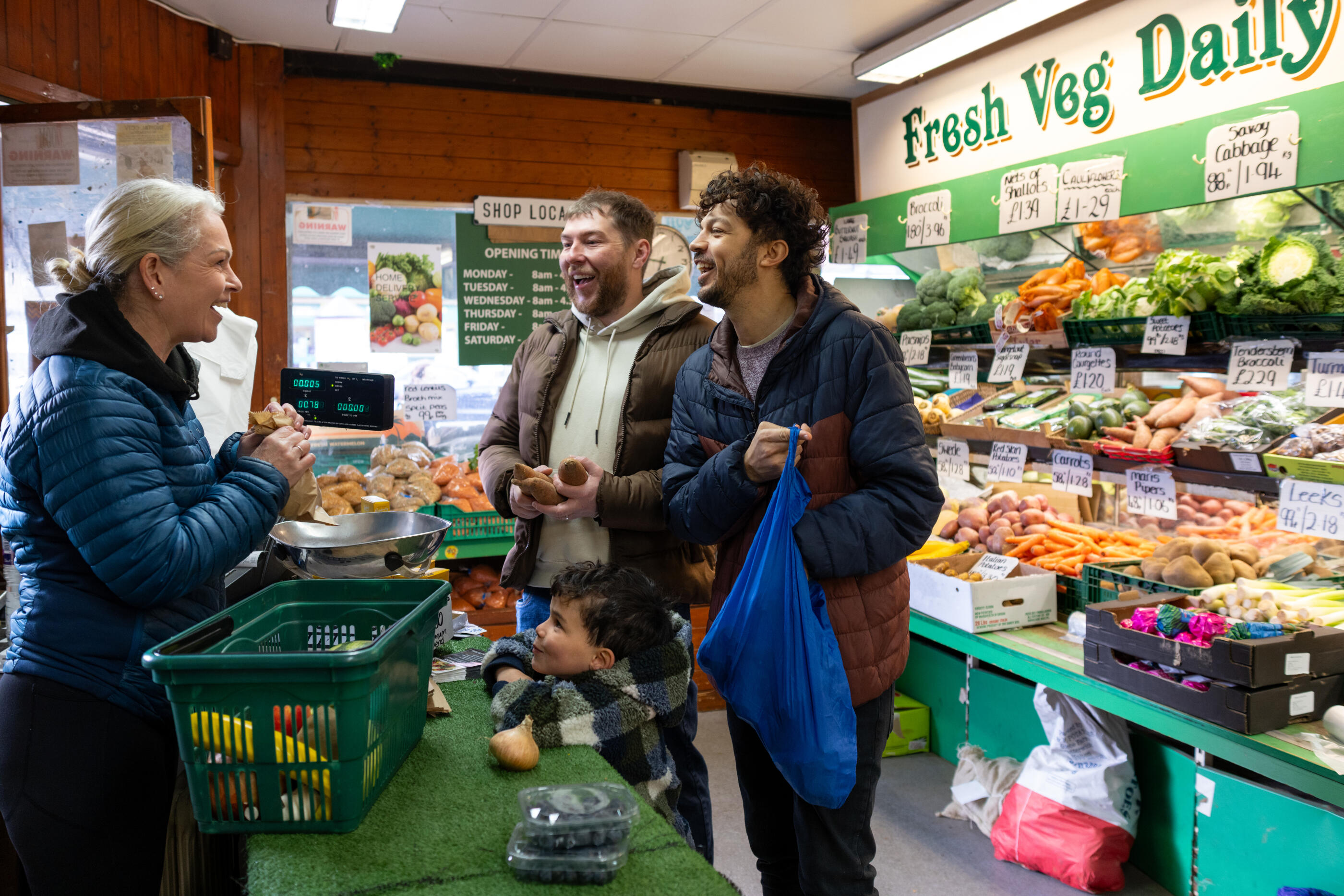 A wide shot of a happy family shopping for fresh produce at a local greengrocer. Two fathers and their child interact with a friendly shopkeeper while buying vegetables. One of the fathers is holding a blue shopping bag while filling it up with the purchased goods.Videos are available similar to this scenario.