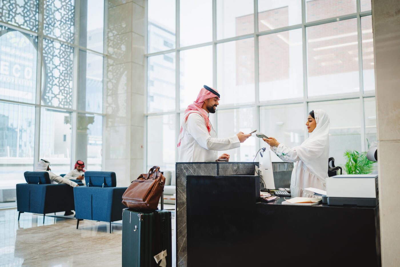 Young Middle Eastern businessman in modest attire making a contactless payment at a modern hotel reception desk, with luggage nearby. large windows and contemporary decor