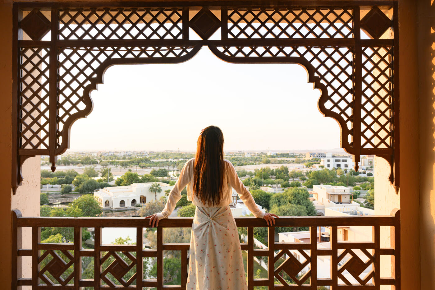 Young woman standing on an ornate wooden balcony, looking out at the sprawling city landscape with lush greenery under a warm, clear sky, embracing relaxation and travel experience