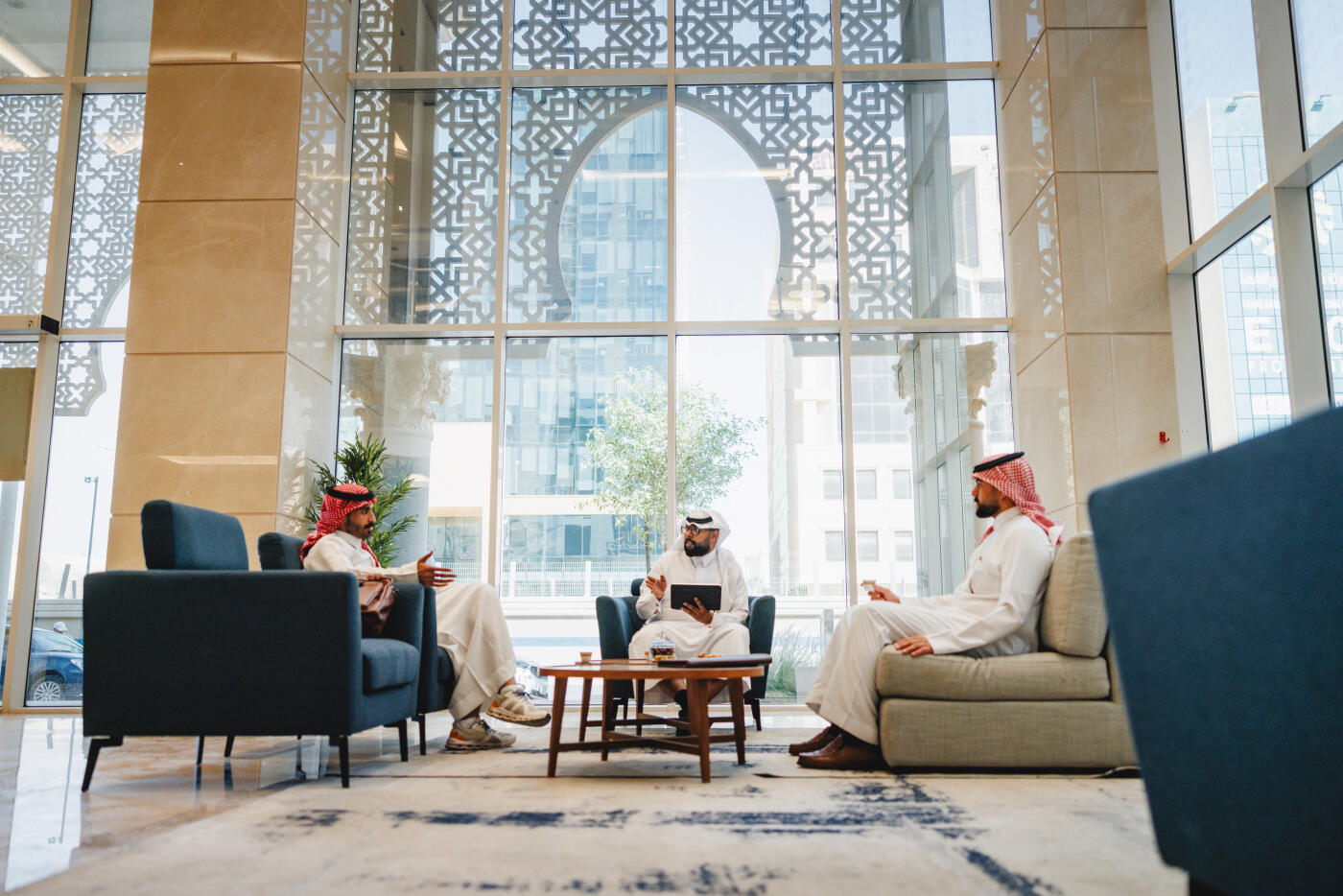 Two Middle Eastern men in business attire engage in discussion within a modern luxury hotel lobby. Natural light streams through geometric patterned screens, illuminating contemporary seating arrangements and a small wooden table between them