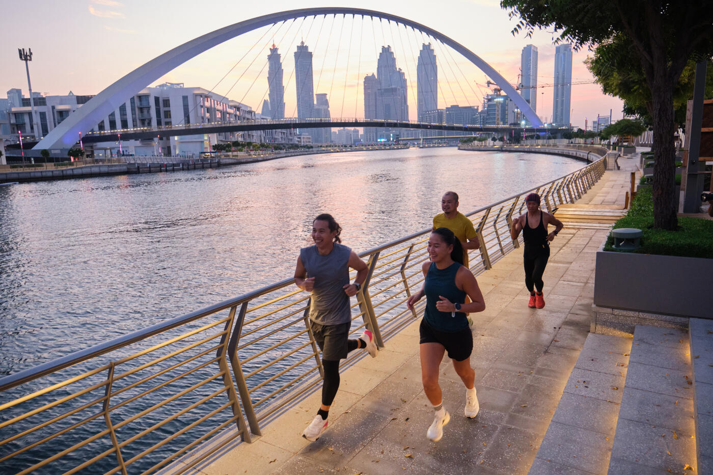 High angle view of happy adult friends jogging on promenade near water canal. People are doing their morning routine. Tolerance bridge can be seen against sky.