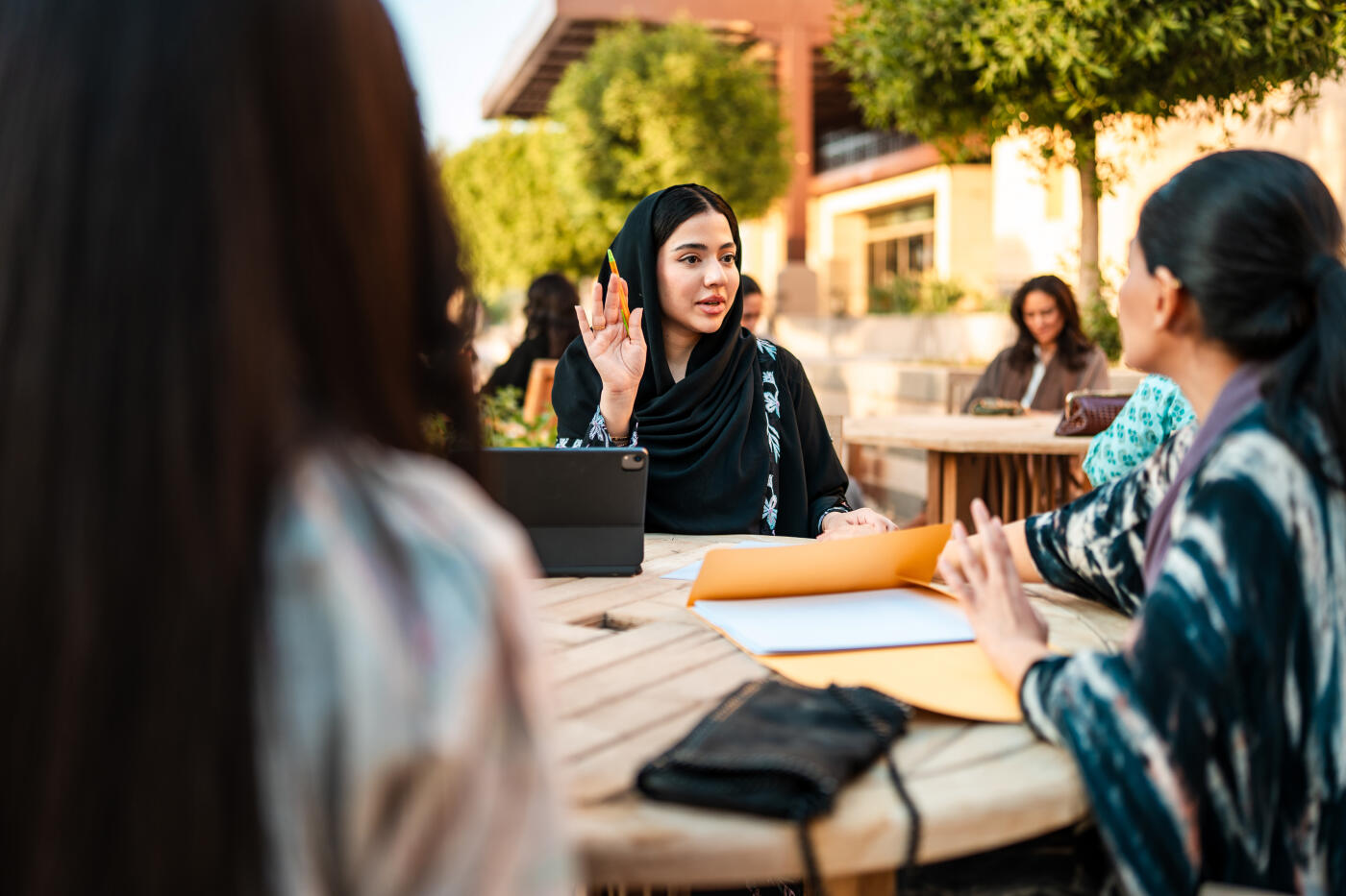 Young Middle Eastern female university students collaboratively discussing academic topics at an outdoor setting with notebooks and a laptop, showing engaged interaction and teamwork.