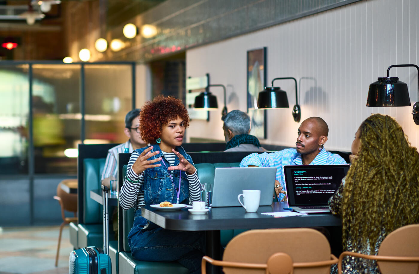 Casual business person with curly hair talking ti two co-workers in meeting, suitcase by table