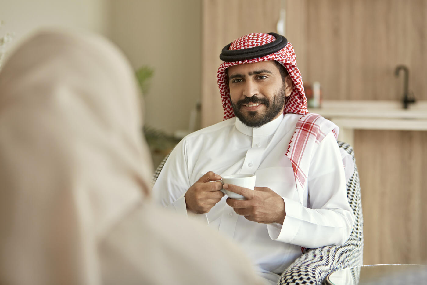 Over the shoulder view of contented man in traditional Saudi attire sitting in domestic room with family and enjoying weekend leisure.