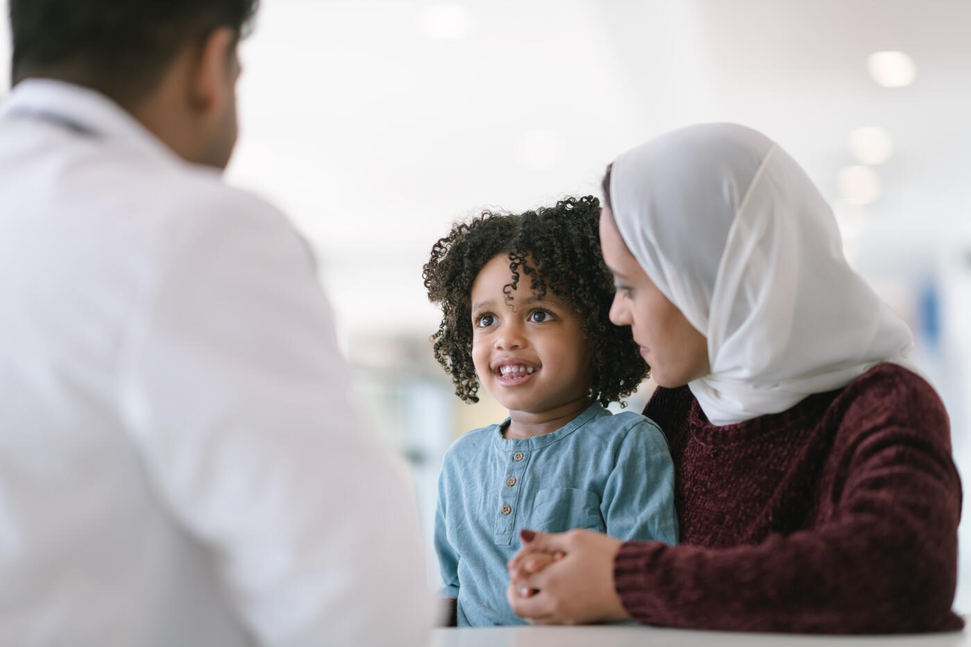 A young Muslim mother wearing a hijab takes her toddler son to visit the pediatrician. The boy is sitting on his mother's lap. They are seated at a table and a male doctor is seated across from them. The child is smiling at the doctor. The mother is looking at her son.