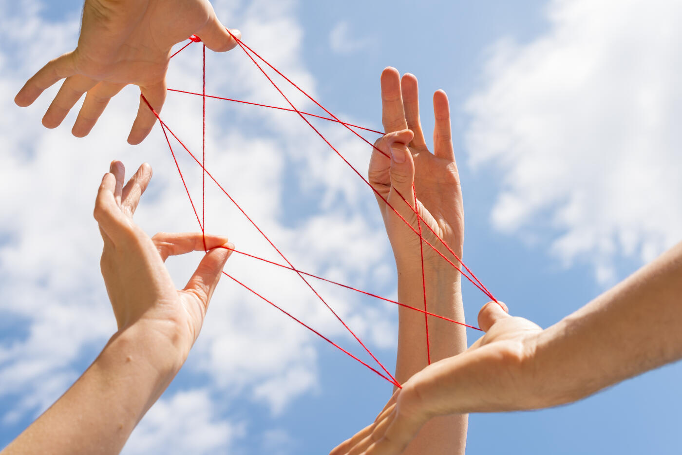 Four hands (female and male) against blue sky playing cats cradle with red wool.