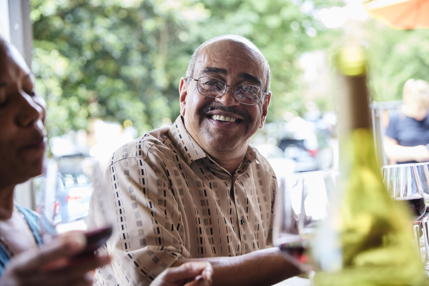Smiling senior man, wearing eyeglasses, engaged in conversation with friends during a wine tasting event at a restaurant.