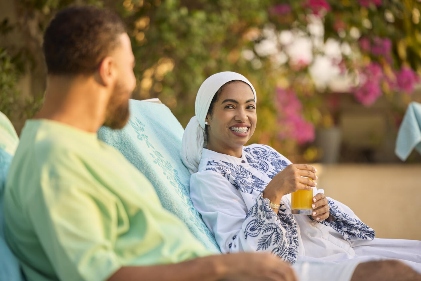 Smiling middle eastern woman spending leisure time talking with man. Arab female is wearing abaya while sitting on deck chair. She is holding juice glass in hand.