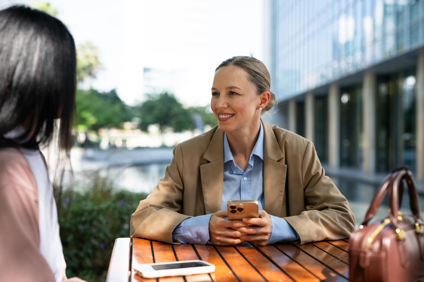 Two businesswomen engaged in conversation at an outdoor table in the Dubai office district. The scene features modern architecture and a tranquil water feature. The women appear focused and professional, with digital devices on the table, under bright daylight.