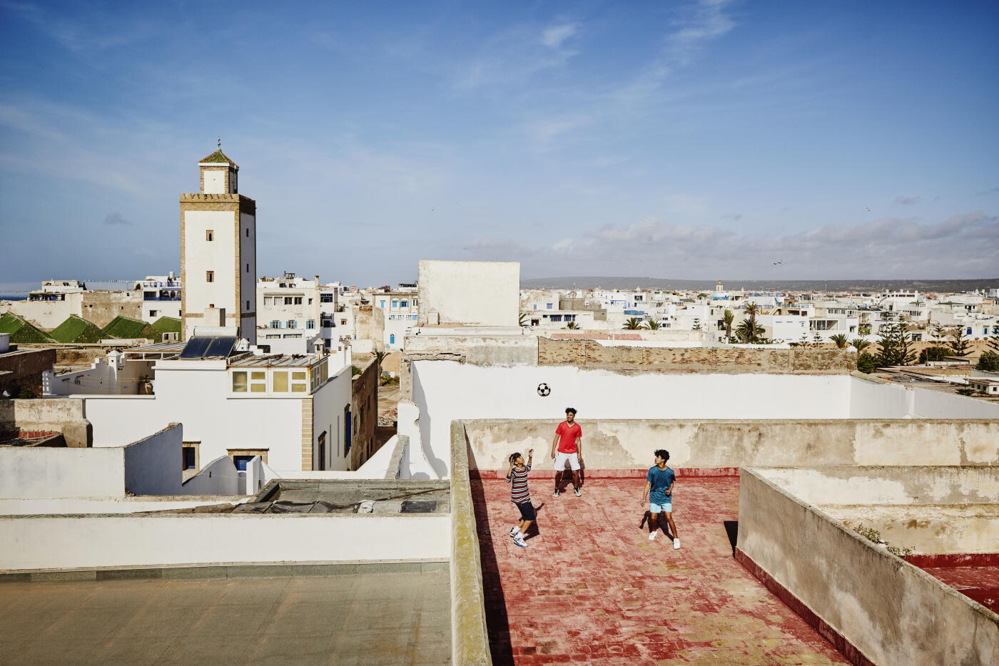 Extreme wide shot high angle view of young male friends juggling soccer ball together on rooftop overlooking Essaouira on sunny afternoon