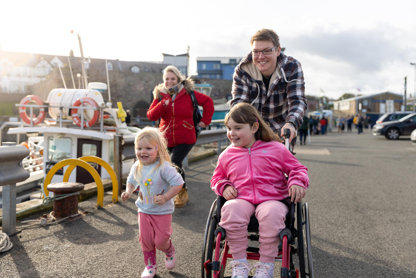 A family with two children enjoying a day out together in Beadnell, North East England. They are moving past the harbour and the main focus is the father running while pushing his daughter who is a wheelchair user. They all look excited.