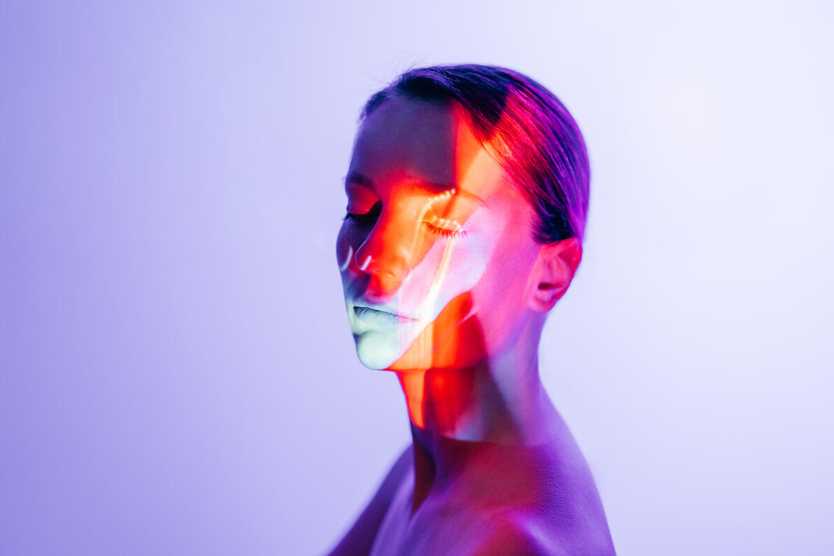 A woman with closed eyes is illuminated by bold red, blue, and violet projections. The conceptual beauty portrait expresses digital transformation, sensory wellness, and the intersection of human identity and technology.