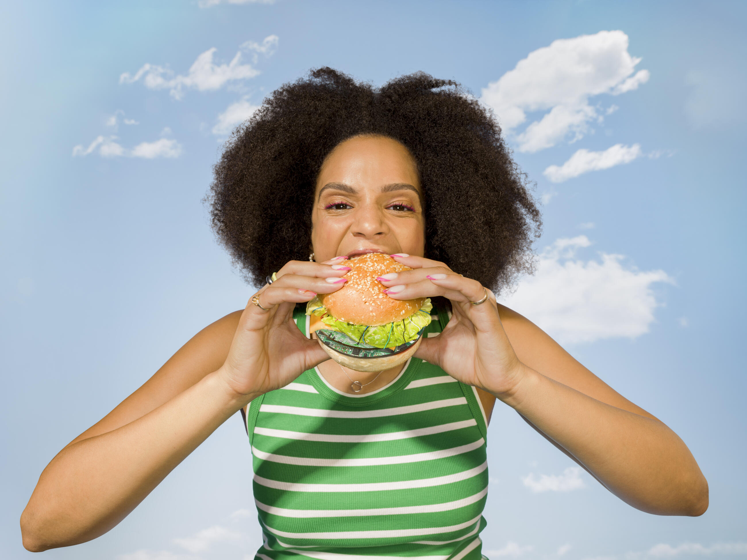 コンピューターケーブルがついた大きな回路基板のハンバーガーを一口食べる若い女性のポートレート