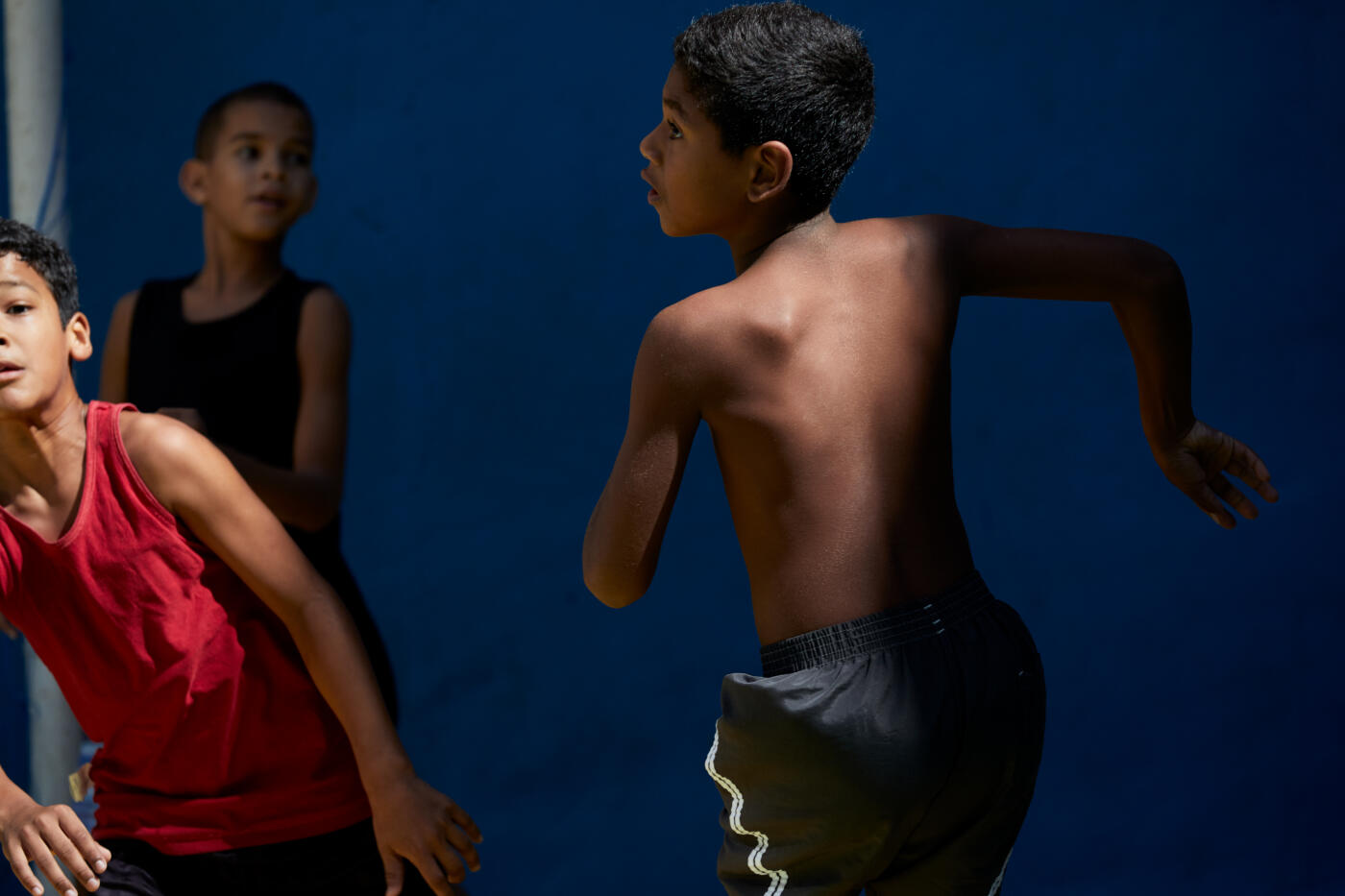 Group of three boys playing football on a brightly coloured court in a favela in Rio de Janeiro. One of the boys is wearing red top, one is wearing a black top and the other is not wearing a shirt.