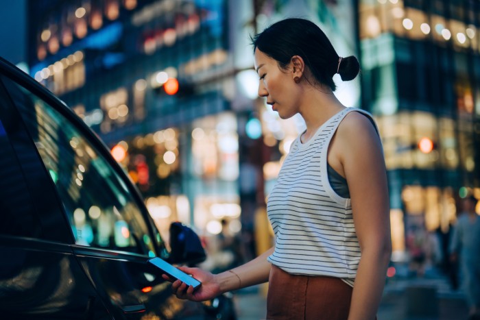 Young Asian woman using mobile app device on smartphone to unlock the doors of her intelligence car in the city at night, against illuminated buildings. Wireless and modern technology. Smart car with digital car key concept