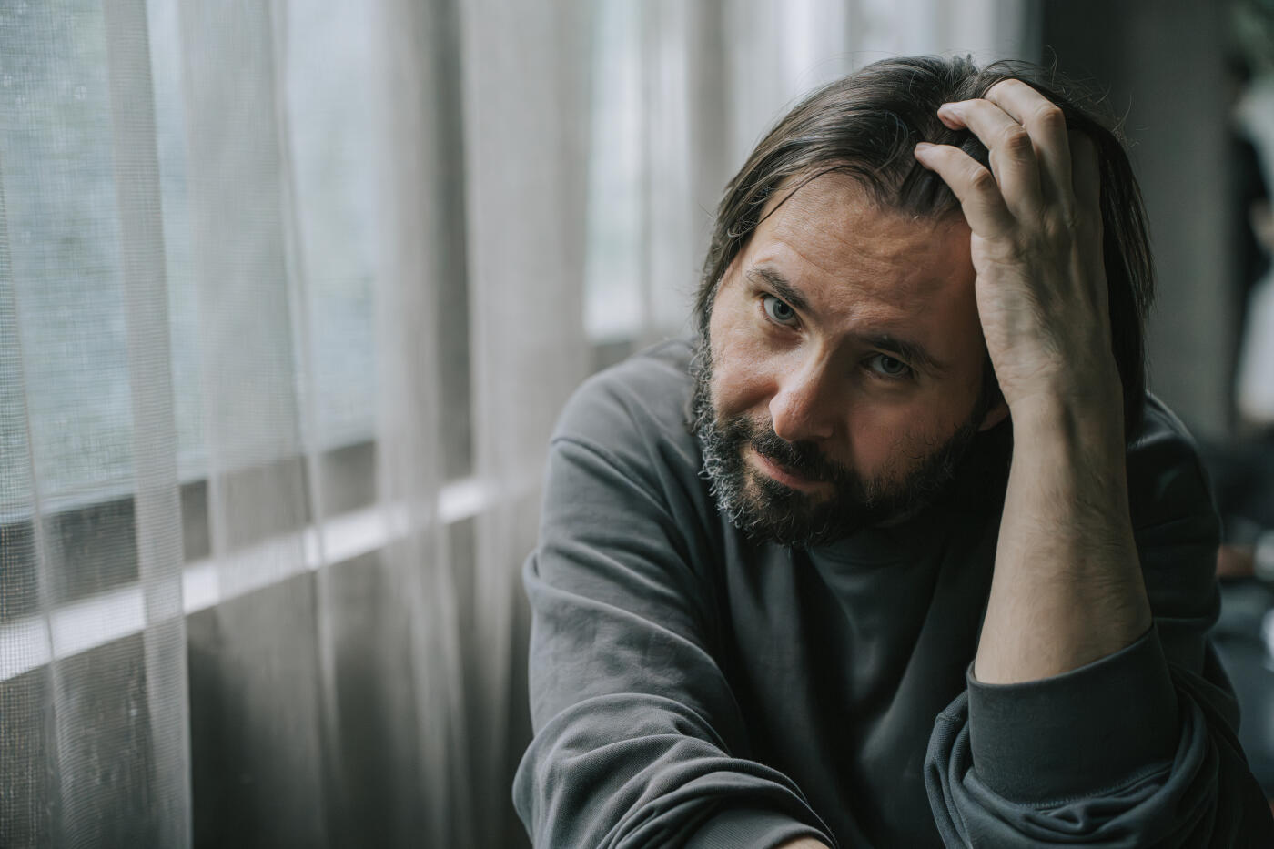 Close-up portrait of a bearded Caucasian man in casual wear, resting his head on one hand while looking intently at the camera. Natural light filters through window curtains, casting a soft, melancholic tone. The mood suggests introspection, mental health awareness, burnout, or emotional fatigue.