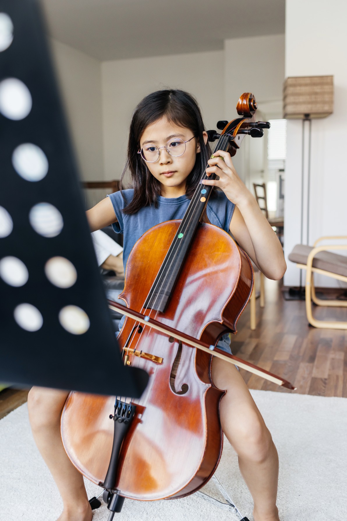 focused young girl sits upright, carefully playing the cello while reading from a music stand. Her expression shows determination and concentration as she perfects her performance. The warm lighting and home setting make this a peaceful and inspiring practice session.