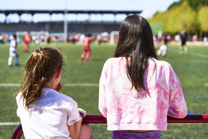 rear view of two girls standing leaning on the metal handrail in casual clothes watching the soccer game.