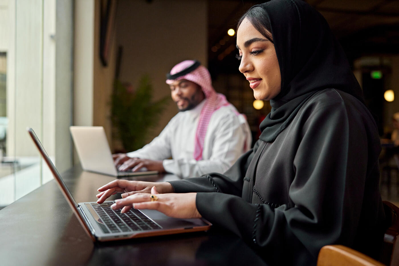 Side view with focus on businesswoman in mid 20s wearing abaya and hijab, sitting at window table and typing, businessman in background doing the same.
