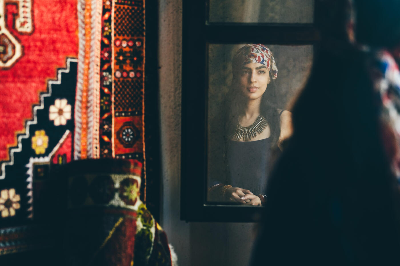 A young woman in a headscarf and necklace is seen reflected in a framed mirror, looking directly at the camera with a calm, serene expression. The image is intimate and culturally rich, conveying a sense of self-reflection and mysterious beauty.