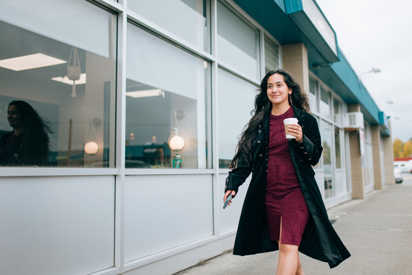 A stylish Indigenous Alaskan woman walks down an Anchorage Alaska street holding a hot cup of coffee and her smartphone.  Headed to the next business deal.