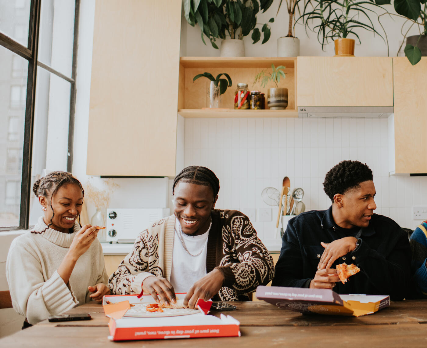 Three young people sit at a dining table, about to tuck in to a fresh, hot, delicious pizza. The scene is relaxed, fun, and chaotic.