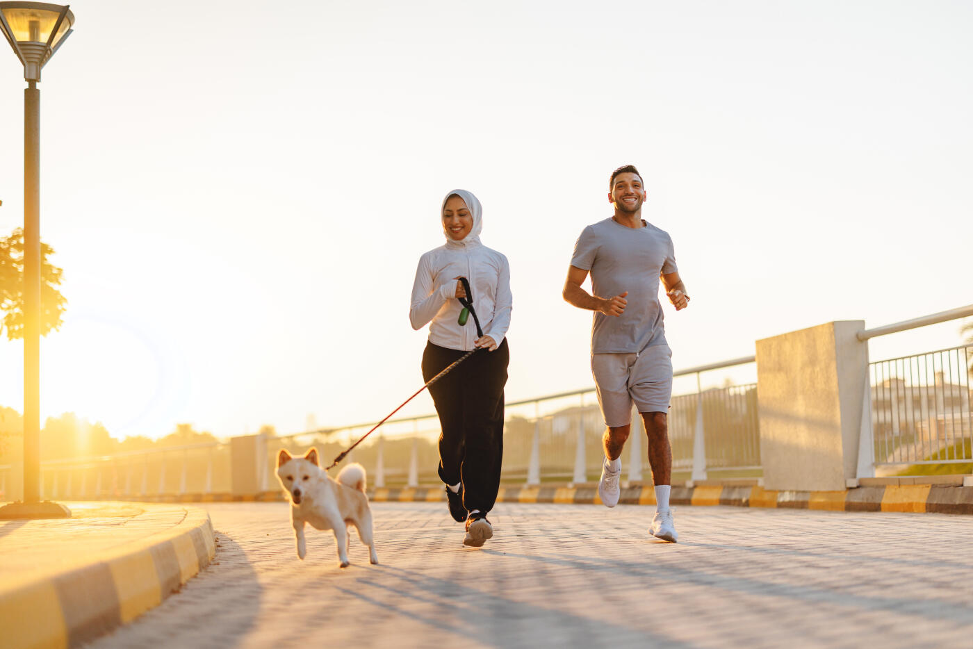 A mid adult Hispanic male and mixed race female jogging together in Dubai during sunrise, accompanied by a small dog on a leash, exuding energy and joy.