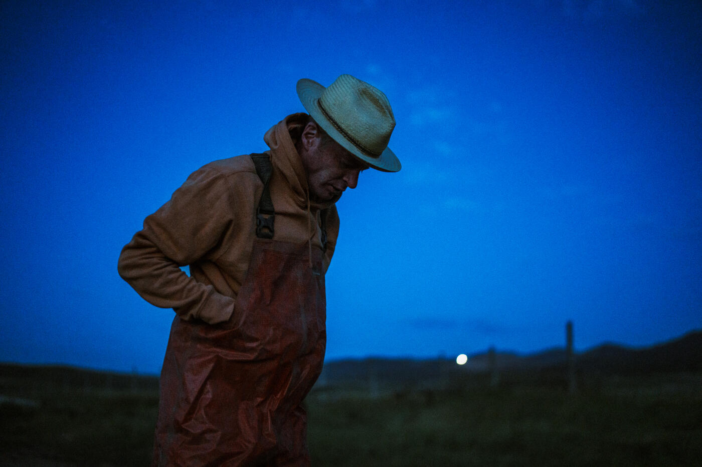 At first light, a rancher readies himself for a day of fieldwork, adjusting his waterproof gear beneath a deep blue Wyoming sky.