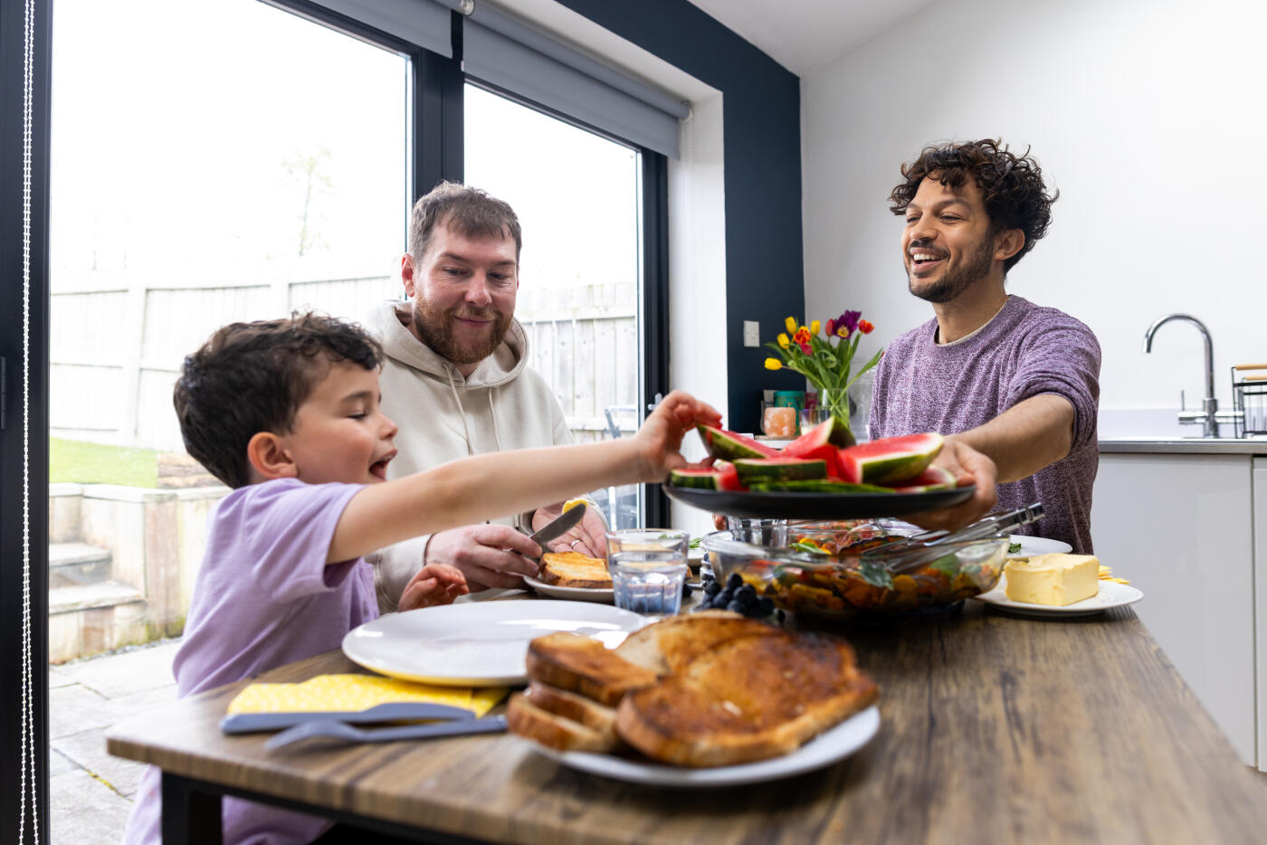 A wide shot of a family having a meal together while sitting at a table in a kitchen in their family home. The two fathers are looking at their son while smiling, and one of them is passing the boy a plate of watermelon slices. An array of food is presented on the table for the family to eat.Videos are available similar to this scenario.