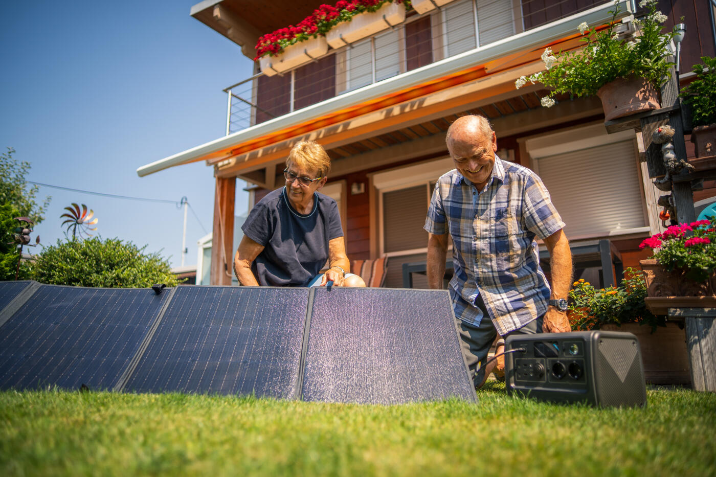 Happy smiling senior couple just set up new portable solar panels with battery outdoors in their garden on sunny summer day, ready for producing some of their power consumption with electricity from sun energy