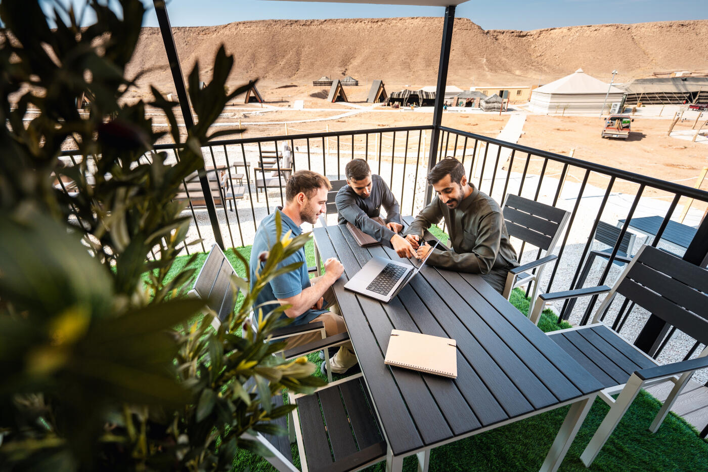 Diverse businessmen collaborating around a laptop on a furnished terrace at Ghamra Desert Camp. The outdoor workspace overlooks sandy desert terrain under bright sunlight, with plants framing the scene and documents spread on the table.