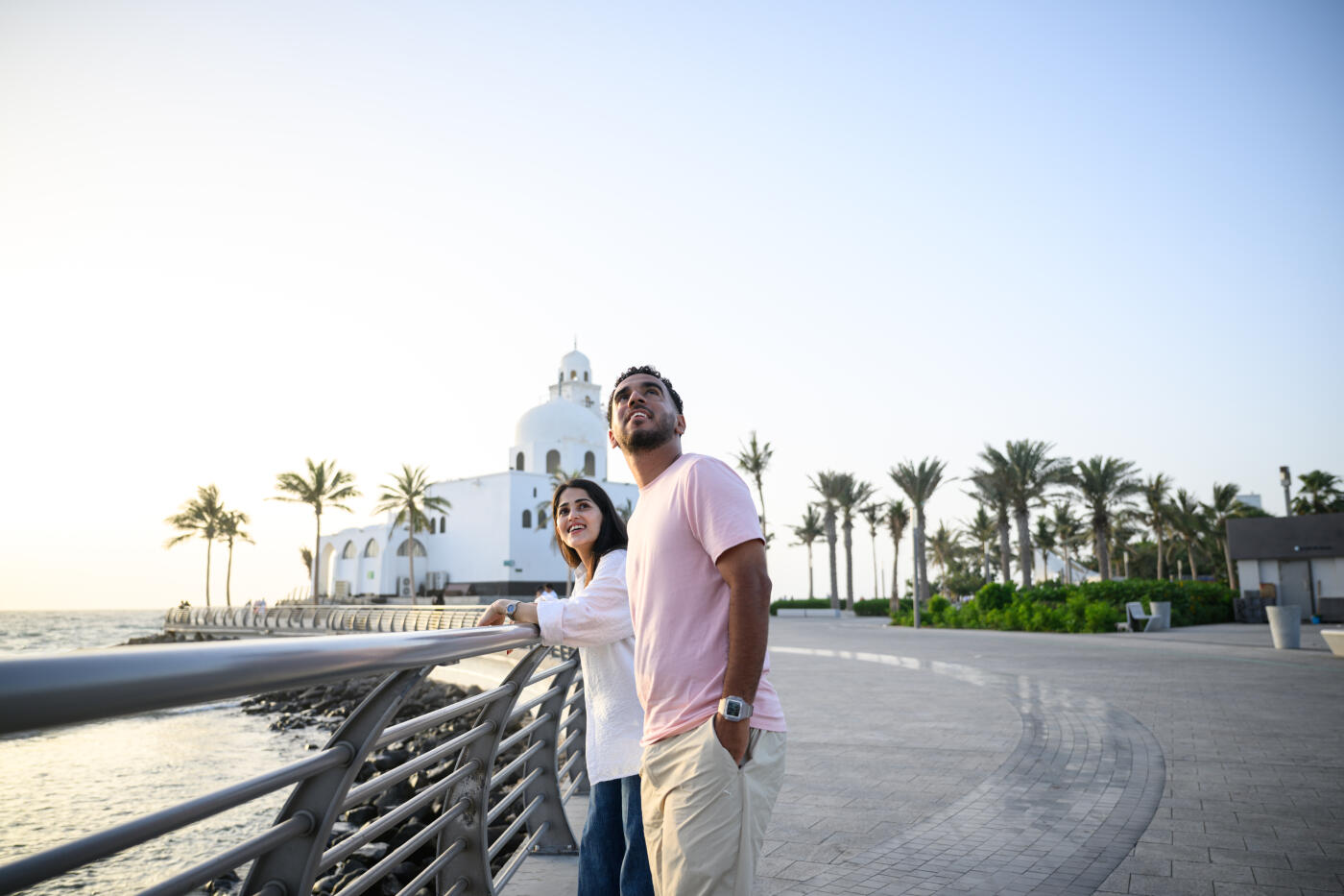 Candid shot of a curious couple standing together by a railing, looking up at the sky with palm trees and white architecture highlighting the Corniche promenade.