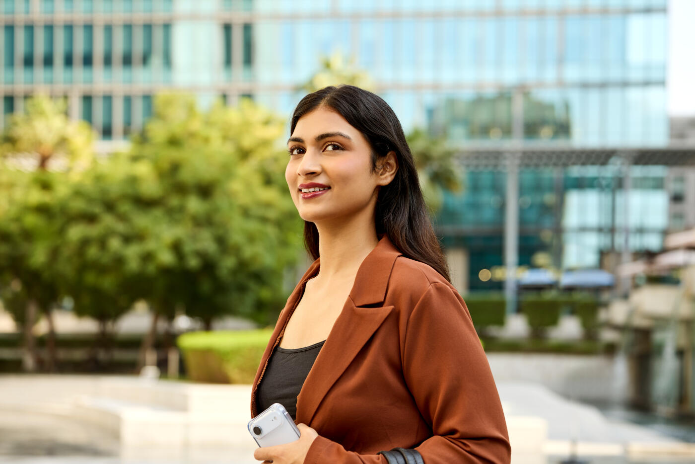 Smiling businesswoman standing outdoors while holding her smart phone. Young entrepreneur dressed in formal pant suit. She is looking away with thoughtful and happy expression.
