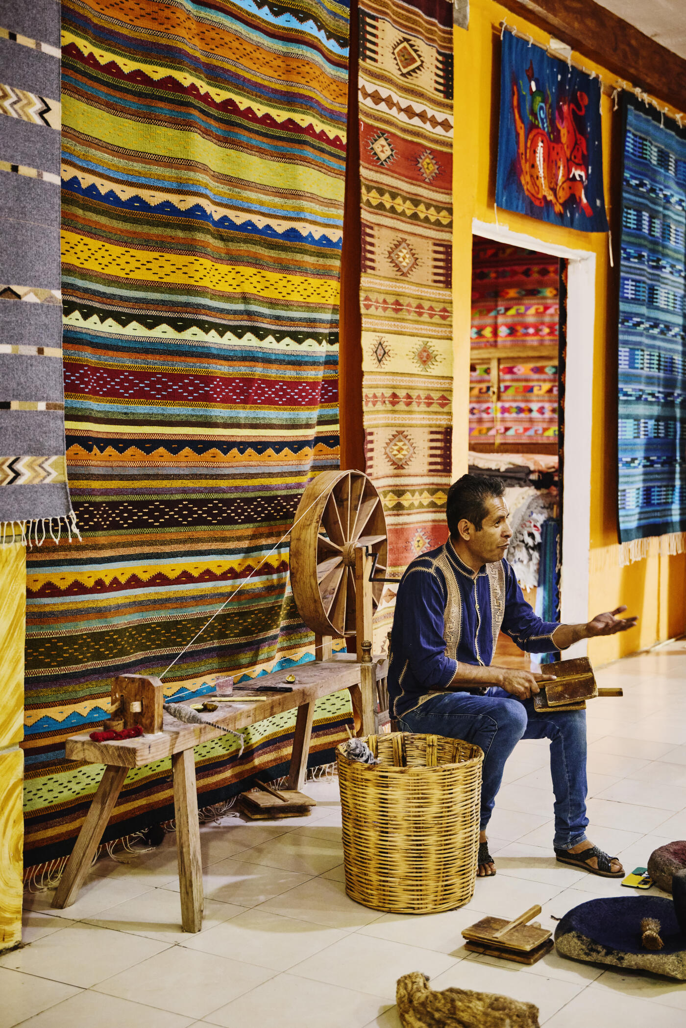 Wide shot of male artisan teaching traditional Mexican fiber arts to students while sitting by spinning wheel and demonstrating with hand cards
