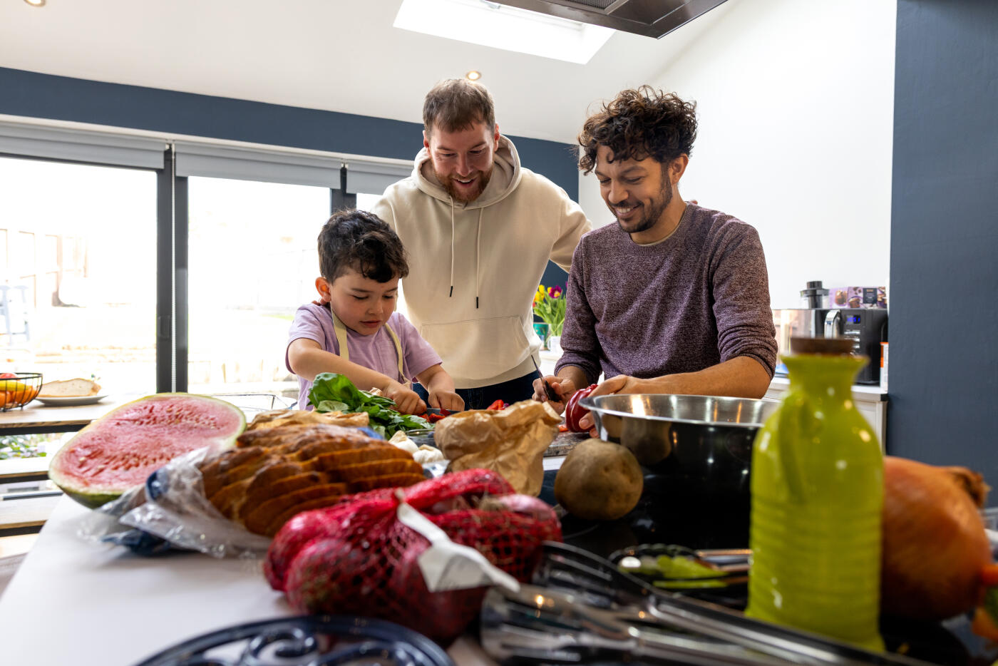 A wide shot of two fathers and their young son preparing fresh vegetables together in a modern kitchen. The child is learning to cut vegetables as his parents watch attentively. They are preparing food for a healthy family meal. A scene of family bonding and a healthy lifestyle at home. A spread of food can be seen on the table in front of them, such as sliced bread and watermelon.Videos are available similar to this scenario.
