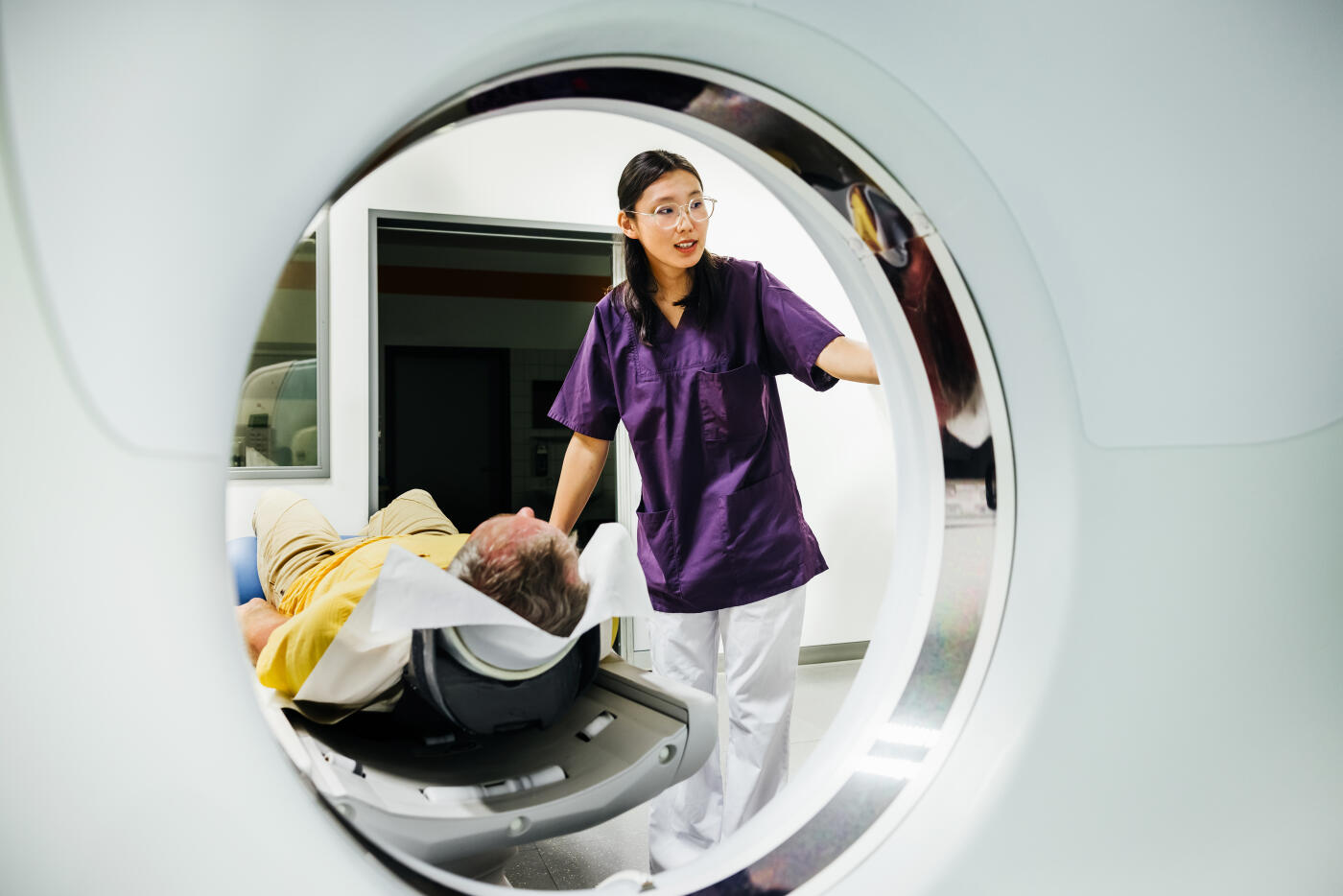 A young female medical technician in a purple uniform prepares a patient for a CT scan inside a hospital. The patient, lying down in the scanner, wears a yellow shirt and beige pants.