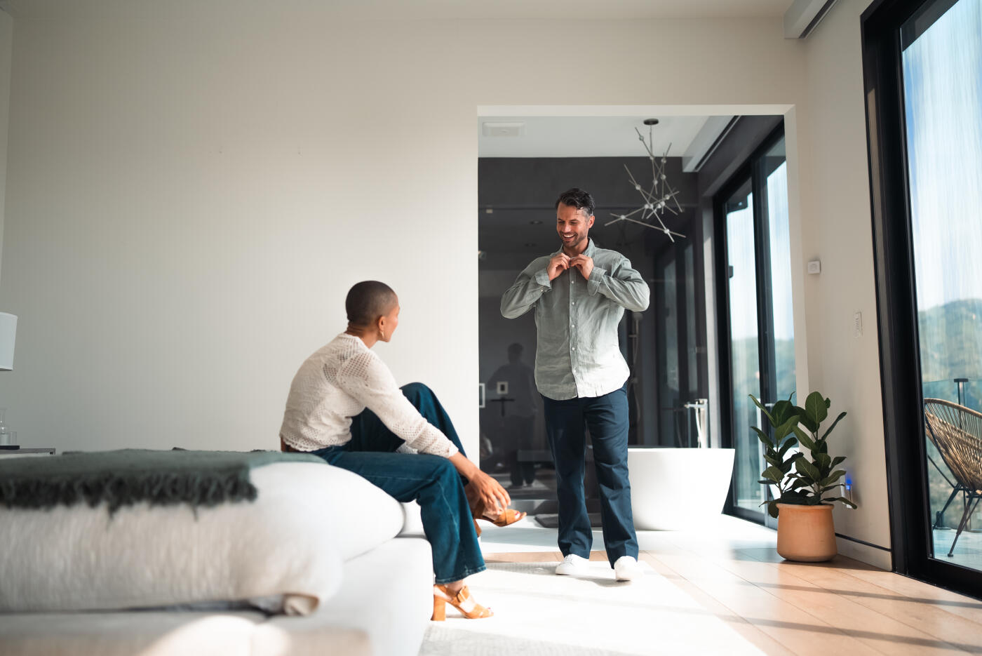An affluent interracial couple gets ready together in their elegant primary bedroom suite, surrounded by sophisticated décor and breathtaking panoramic mountain views.