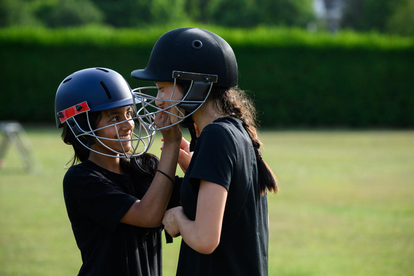 Teenage girls laughing and helping each other kit up with protective helmets for cricket practice