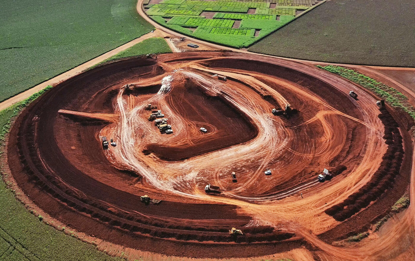 Aerial image taken during the day over a large construction site in a rural area of Brazil, showing land movement and heavy machinery operating on red soil. The site is surrounded by farmland, and several trucks and excavators are visible. No people can be individually identified. The clear sky and bright lighting highlight the scale and structure of the earthwork in progress.