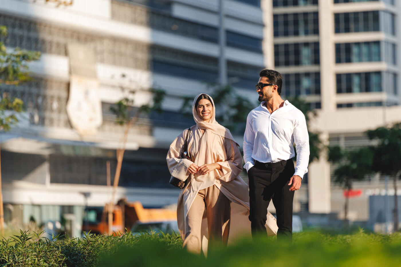 A young Middle Eastern woman wearing a beige hijab and abaya and a Caucasian man in a white shirt walk outdoors in Dubai?s modern urban landscape. The sunny setting highlights contemporary architecture and greenery.